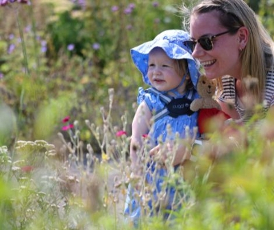 Woman with blonde hair wearing sunglasses kneeling down cuddling a little girl in a blue dress holding a teddy bear looking at flowers.
