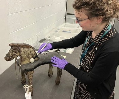 Lady with purple rubber gloves holding a brush delicately wiping an old wooden toy horse.